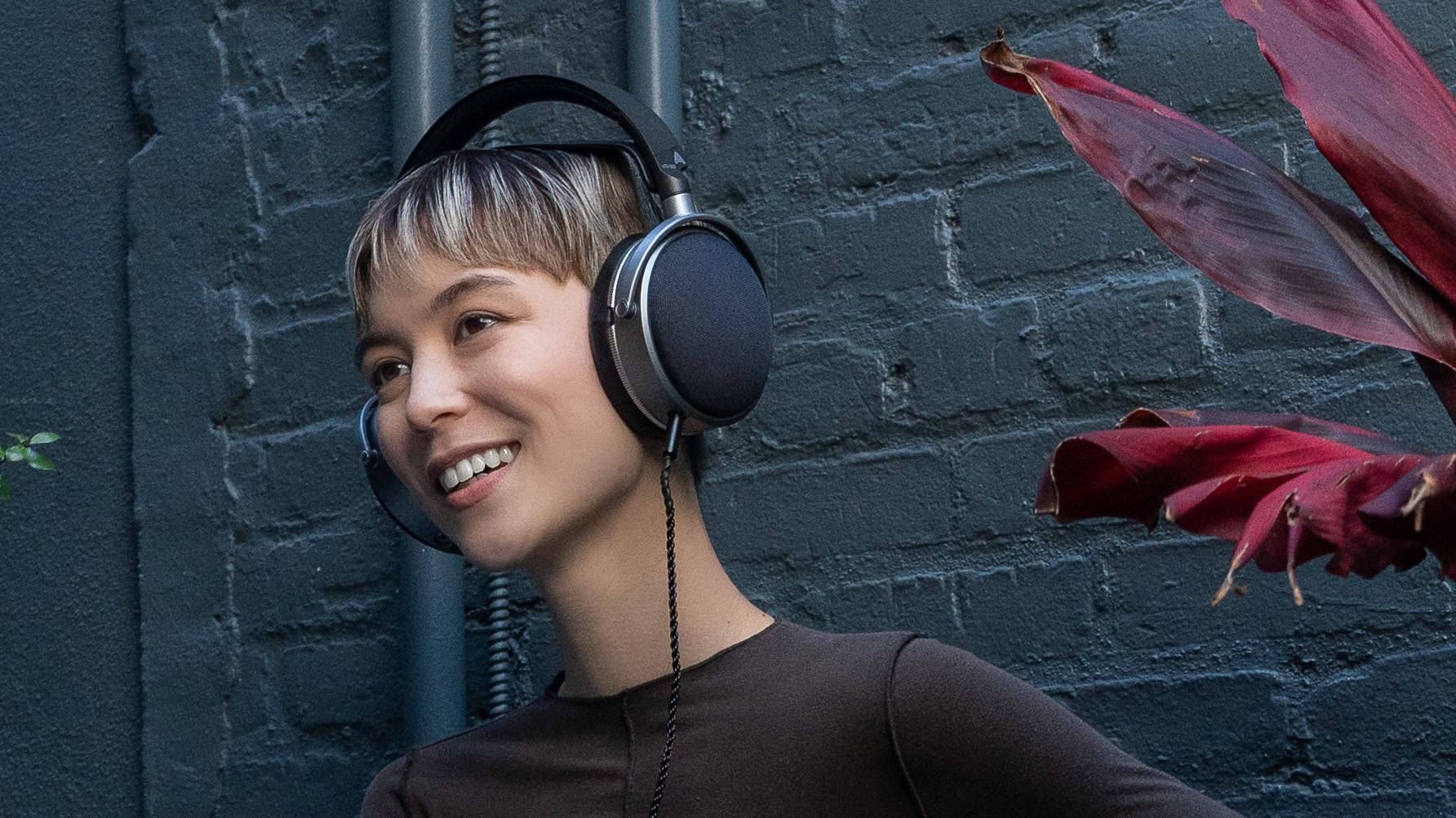 A young androgynous-looking person wearing Audeze LCD-S20 planar magnetic headphones in front of a dark grey brick wall and a dark red plant that reminds me of a Triffid. 